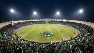 Packed Pakistani cricket stadium at night with floodlights and fans waving green flags